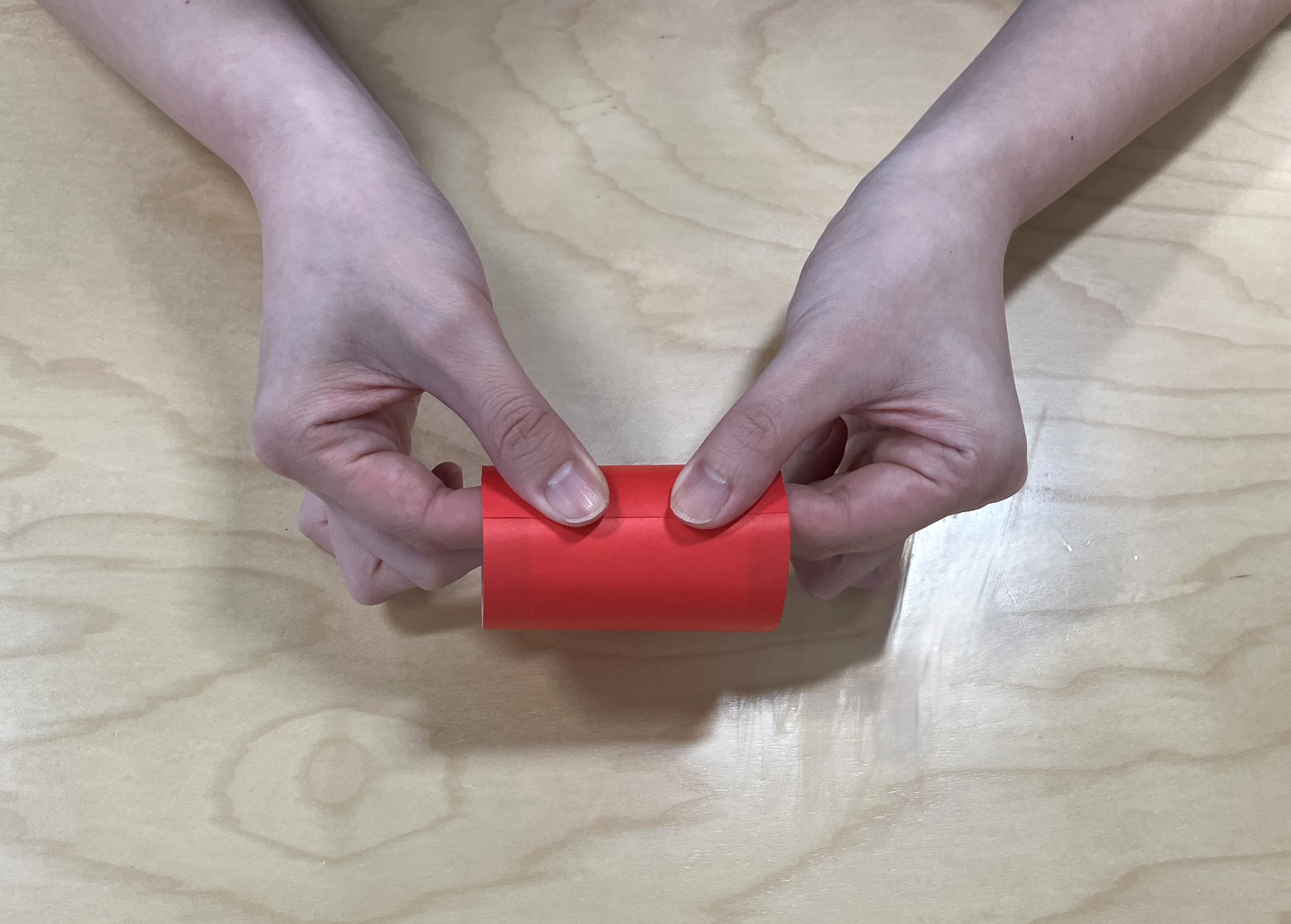 2. 半分に切った折り紙をトイレットペーパーの芯に巻き、巻き終わりをスティックのりでとめて、両端を内側に折り込みます